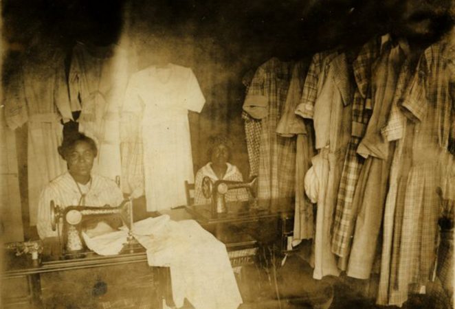 black and white image of two Black women sitting a sewing machines surrounded by dresses