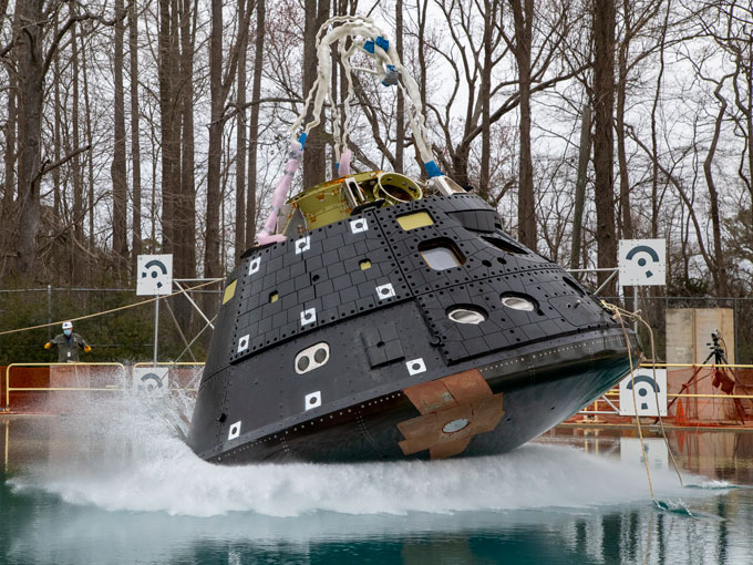 The Orion crew capsule, a cylindrical apparatus, being dropped into a water-filled basin with a forest in the background